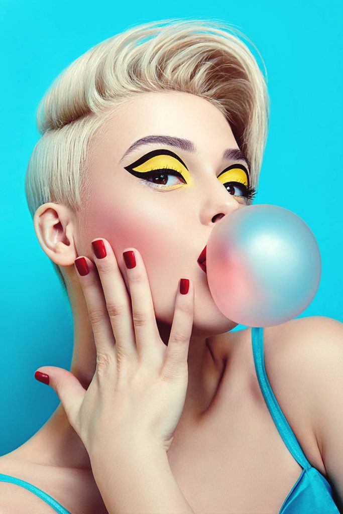 A beautiful young woman with brightly colored makeup and styled hair blowing bubblegum