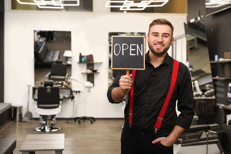 A handsome young barber holding an "Open" sign in his barber shop